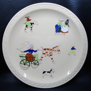 May include: A cream-colored ceramic plate with a scalloped rim featuring hand-painted illustrations of figures in traditional attire. The scenes include a person riding a goat, a person pulling a cart with a donkey, and a small black and white dog.