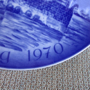 May include: Blue and white ceramic plate with a scene of a bridge and water. The plate has the year "1970" and the word "dag" written on it.