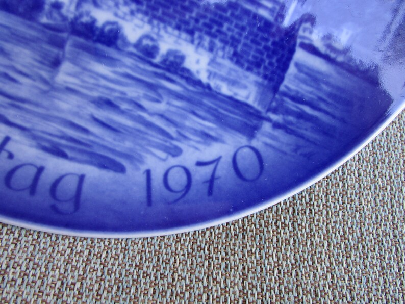 May include: Blue and white ceramic plate with a scene of a bridge and water. The plate has the year "1970" and the word "dag" written on it.