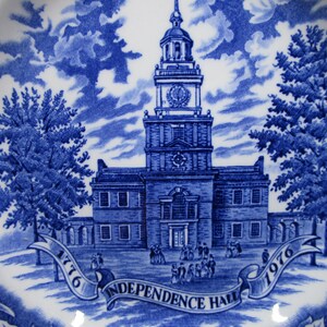 K&ouml;nnte beinhalten: Blau-wei&szlig;e Keramikplatte mit detaillierter Illustration der Independence Hall, mit den Jahren 1776 und 1976 auf einem Banner. Das Design umfasst B&auml;ume, Wolken und architektonische Details.