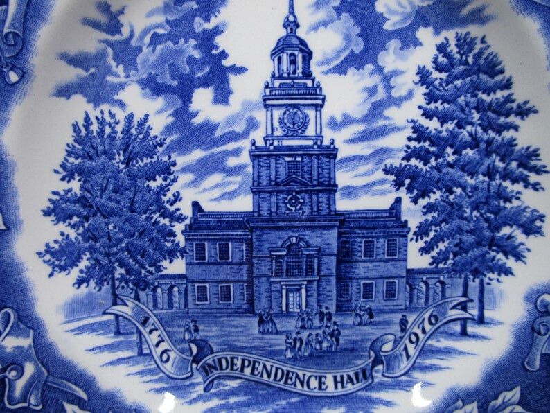 K&ouml;nnte beinhalten: Blau-wei&szlig;e Keramikplatte mit detaillierter Illustration der Independence Hall, mit den Jahren 1776 und 1976 auf einem Banner. Das Design umfasst B&auml;ume, Wolken und architektonische Details.