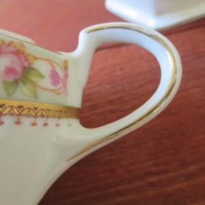 May include: A white porcelain creamer with a gold trim and floral design. The handle is also trimmed in gold.