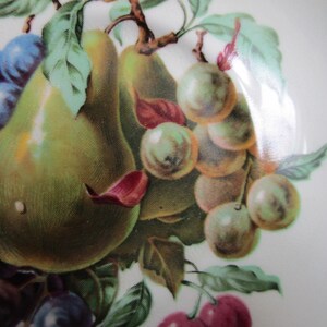 May include: A vintage ceramic plate with a hand-painted design of pears, grapes, and cherries. The pears are green and yellow, the grapes are purple and blue, and the cherries are red.