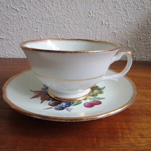 May include: A white teacup and saucer with a gold rim and a floral design featuring cherries and grapes.