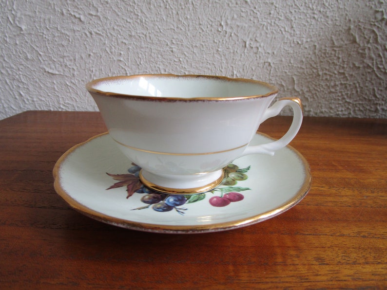 May include: A white teacup and saucer with a gold rim and a floral design featuring cherries and grapes.