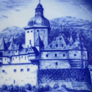 May include: A blue and white ceramic plate with a detailed illustration of a castle in a mountainous landscape. The castle has a tall tower and multiple smaller towers, and the landscape is depicted with a blue and white wash.