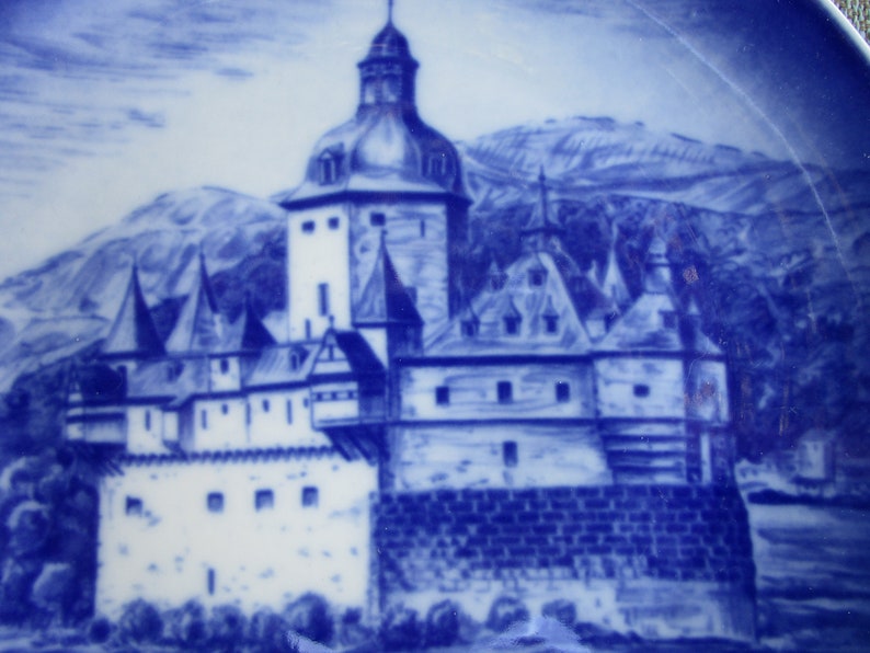 May include: A blue and white ceramic plate with a detailed illustration of a castle in a mountainous landscape. The castle has a tall tower and multiple smaller towers, and the landscape is depicted with a blue and white wash.