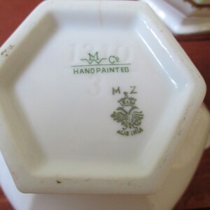 May include: A white hexagonal porcelain teapot base with green markings. The base has the text "M Co. HAND PAINTED" and a green crest with the text "M.Z. AUS IRIA".