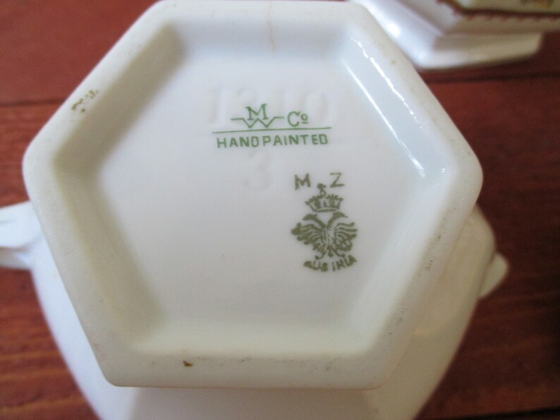May include: A white hexagonal porcelain teapot base with green markings. The base has the text "M Co. HAND PAINTED" and a green crest with the text "M.Z. AUS IRIA".