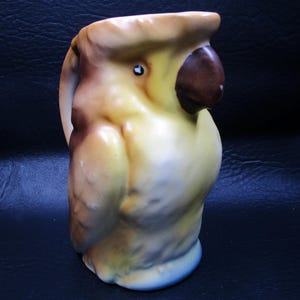 May include: A ceramic pitcher shaped like a parrot, featuring a brown beak and eye, with a yellow, tan, and blue color scheme. The handle is on the left side. The pitcher is a decorative item.