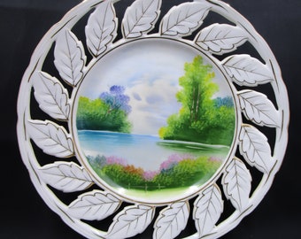 Puede incluir: Plato de cerámica blanco con borde dorado y un diseño de hojas recortadas. El centro del plato presenta una escena de paisaje pintada a mano de un lago con árboles y flores.