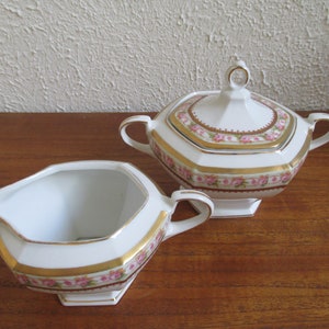 May include: A white porcelain creamer and sugar bowl set with a pink and gold floral design. The creamer and sugar bowl have a octagonal shape with a gold trim.
