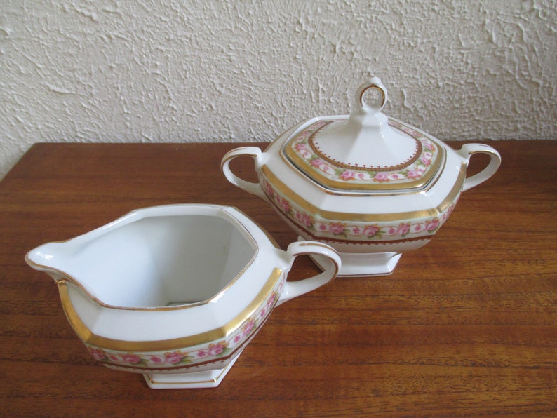 May include: A white porcelain creamer and sugar bowl set with a pink and gold floral design. The creamer and sugar bowl have a octagonal shape with a gold trim.