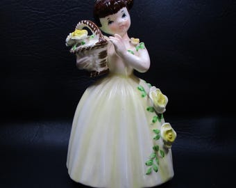 Może przedstawiać: Vintage porcelanowa figurka młodej kobiety w żółtej sukni, trzymającej kosz żółtych kwiatów. Figurka ma brązowe włosy i jest ozdobiona kwiatowymi akcentami. Suknia ma pełną spódnicę i jest ozdobiona żółtymi różami i zielonymi liśćmi.