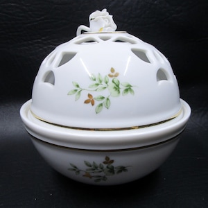 May include: A white porcelain decorative bowl with a domed lid featuring cut-out designs. The lid is topped with a small white rose and accented with gold trim. The bowl is decorated with green and gold floral patterns.