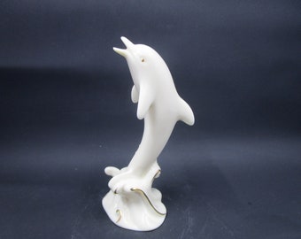 May include: A white porcelain figurine of a dolphin leaping out of the water. The dolphin is painted with gold accents and is set on a base that resembles a wave.
