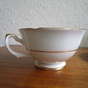May include: A white teacup with a gold rim and a gold band around the middle. The teacup is sitting on a wooden surface.