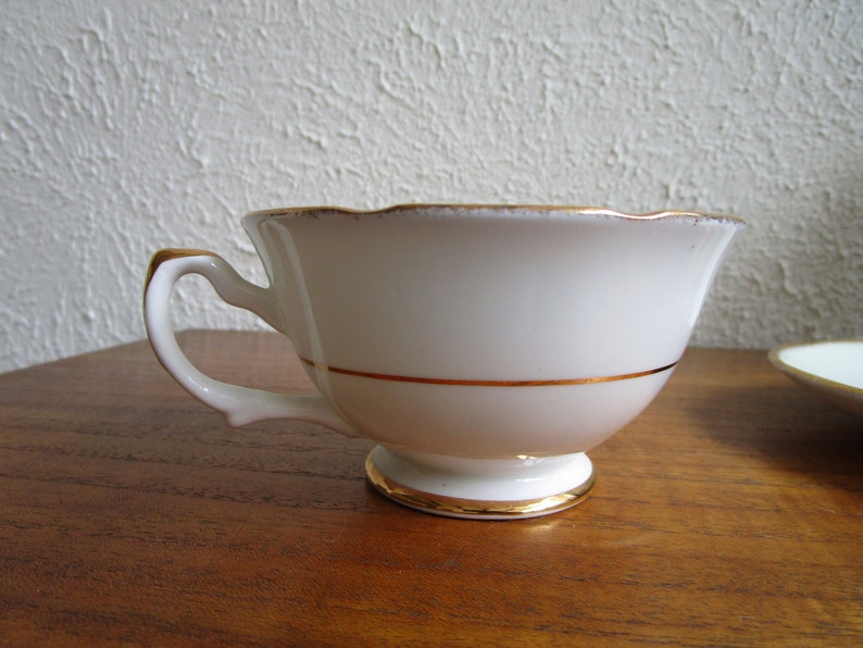 May include: A white teacup with a gold rim and a gold band around the middle. The teacup is sitting on a wooden surface.