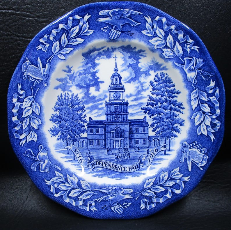 K&ouml;nnte beinhalten: Blau-wei&szlig;e Zierteller mit dem Independence Hall-Motiv. Der Teller zeigt eine detaillierte Illustration des Geb&auml;udes, der B&auml;ume und des Himmels. Der Rand hat ein Blumen- und Adlerdesign. Der Teller ist mit den Jahren 1776 und 1976 gekennzeichnet.