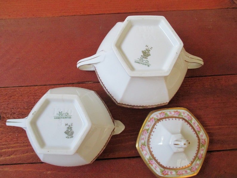 May include: Three white porcelain sugar bowls with gold trim. The bowls are octagonal in shape and have a floral design. The bottom of the bowls have a green and gold logo that reads "M.Z. Co. Hand Painted".