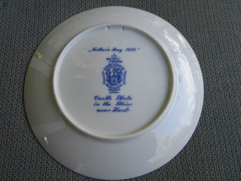 May include: A white ceramic plate with blue text and a blue crest. The text reads "Father's Day 1970" and "Castle Pfalz in the Rhine near Kaub."