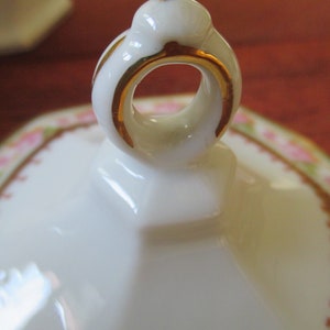 May include: A white porcelain lid with a gold trim and a ring handle. The lid has a floral pattern with pink roses and green leaves.