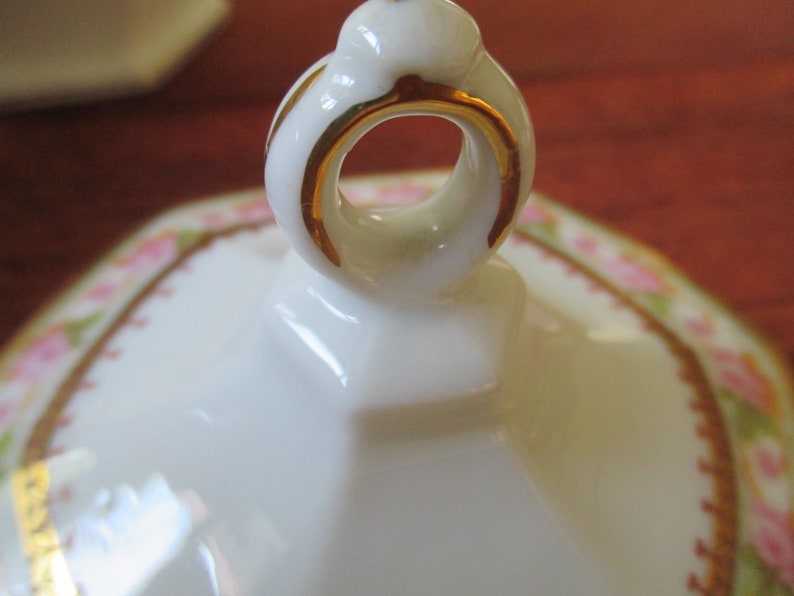 May include: A white porcelain lid with a gold trim and a ring handle. The lid has a floral pattern with pink roses and green leaves.