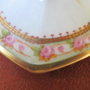 May include: Close-up of a white porcelain plate with a gold and red rim. The plate is decorated with a floral pattern of pink roses and green leaves.