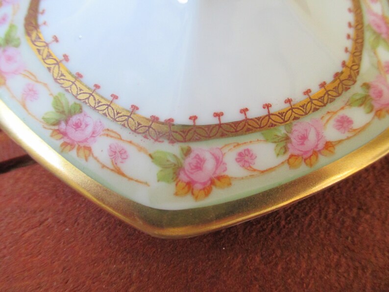 May include: Close-up of a white porcelain plate with a gold and red rim. The plate is decorated with a floral pattern of pink roses and green leaves.