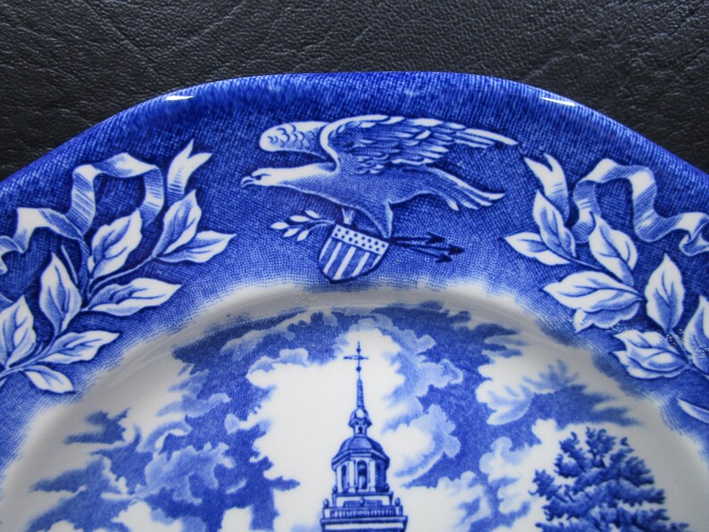 K&ouml;nnte beinhalten: Blaue und wei&szlig;e Zierteller mit einem Adler, Schild und Pfeilen sowie einem Kirchturm und Bl&auml;ttern. Der Teller hat ein detailliertes Design und eine klassische &Auml;sthetik, ideal zum Ausstellen oder Essen.