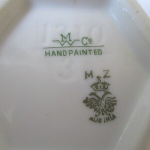 May include: Close-up of a green mark on a white porcelain item. The mark reads "M Co. Hand Painted" and "MZ" with a crown and a bird.