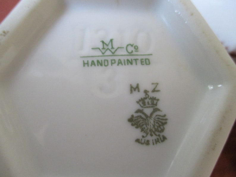 May include: Close-up of a green mark on a white porcelain item. The mark reads "M Co. Hand Painted" and "MZ" with a crown and a bird.