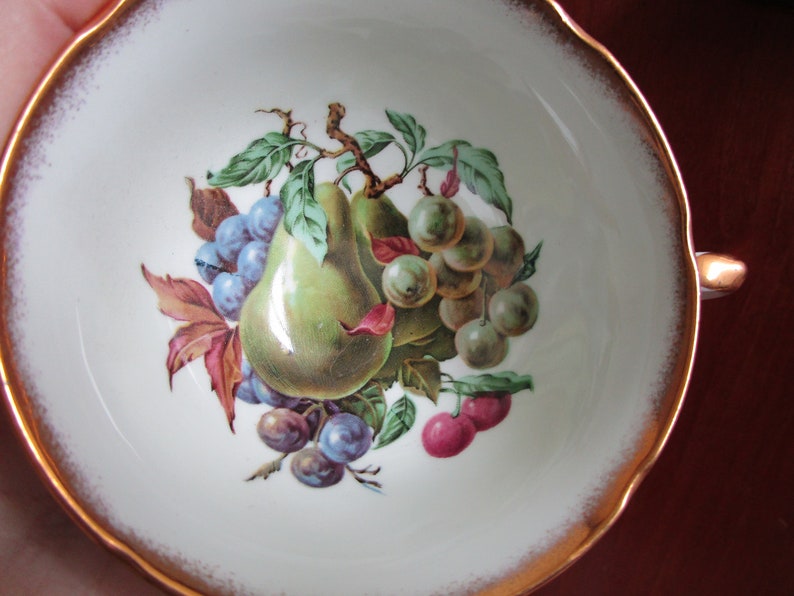 May include: A white porcelain teacup with a gold rim and a painted design of pears, grapes, and plums.