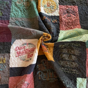 May include: A patchwork quilt composed of various fabric squares, featuring band logos and tour dates. The quilt showcases a mix of colors, including blue, green, red, and black. The text "Dave Matthews Band" is visible on one square.
