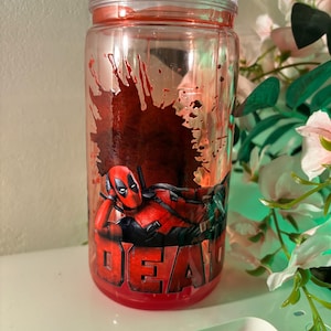 K&ouml;nnte beinhalten: Ein zylindrischer Trinkbecher aus klarem Kunststoff mit einer Grafik der Figur Deadpool in liegender Pose. Das Design zeigt einen dunkelroten Spritzfarbeneffekt hinter der Figur und das Wort DEADPOOL in fetten roten Buchstaben am Boden.