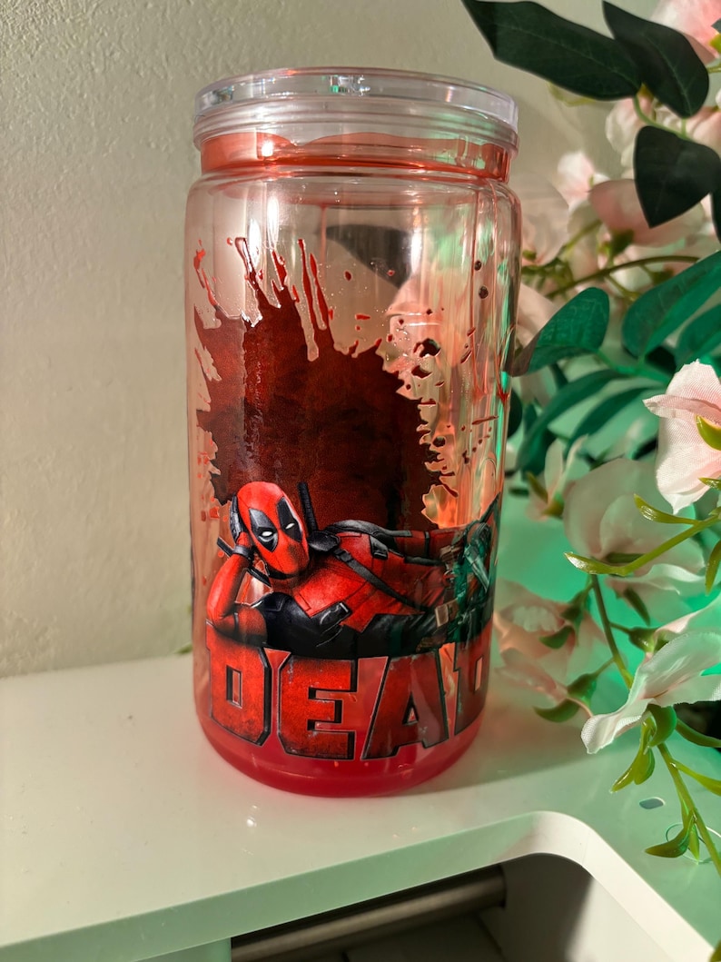 K&ouml;nnte beinhalten: Ein zylindrischer Trinkbecher aus klarem Kunststoff mit einer Grafik der Figur Deadpool in liegender Pose. Das Design zeigt einen dunkelroten Spritzfarbeneffekt hinter der Figur und das Wort DEADPOOL in fetten roten Buchstaben am Boden.