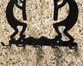 May include: Black metal Kokopelli key holder with five hooks. Two keychains hang from the hooks: a black and white cow and a red and white owl. The key holder is mounted on a speckled wall.