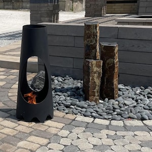 May include: A black metal fire pit with a rounded opening and a decorative base. The fire pit is surrounded by gray stones and is in front of a wall made of gray bricks.