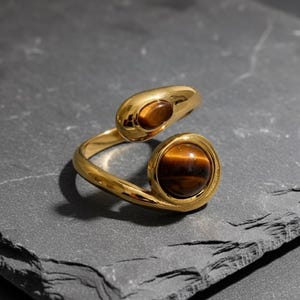 Könnte beinhalten: Ein goldfarbener Ring mit zwei Tigeraugensteinen. Der Ring hat ein einzigartiges, offenes Design mit einem geschwungenen Band, das die Steine hält. Die Steine sind rund und haben eine warme, braune Farbe. Der Ring wird auf einer dunkelgrauen Oberfläche präsentiert.
