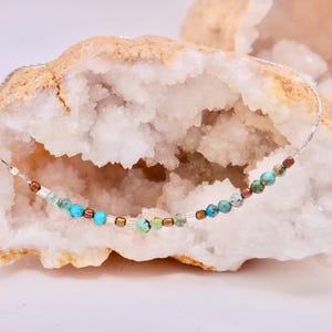 Peut inclure: Un collier délicat en chaîne argentée avec une rangée de perles colorées turquoise, vertes et brunes. Le collier est présenté sur une géode de cristal blanc.