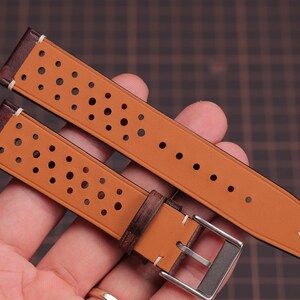 Puede incluir: Correa de reloj de dos piezas en color marr&oacute;n claro con un dise&ntilde;o perforado. La correa tiene detalles en cuero marr&oacute;n oscuro y una hebilla plateada. La correa del reloj se sostiene en una mano.