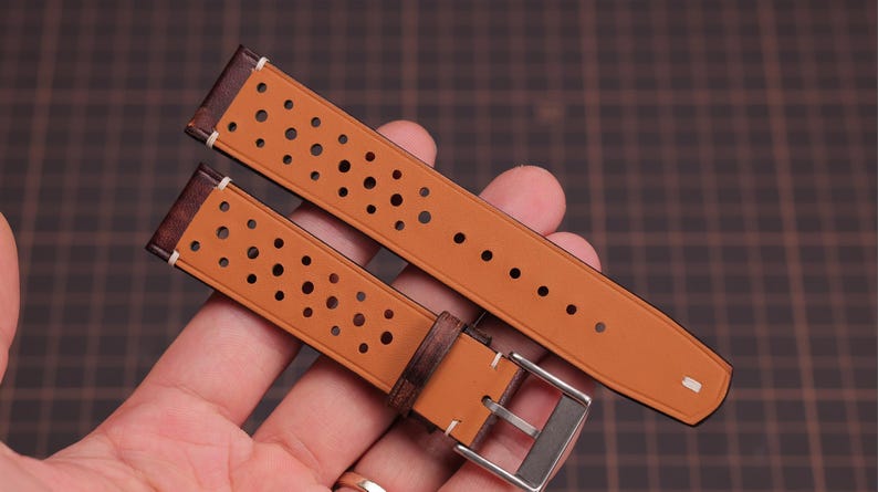 Puede incluir: Correa de reloj de dos piezas en color marr&oacute;n claro con un dise&ntilde;o perforado. La correa tiene detalles en cuero marr&oacute;n oscuro y una hebilla plateada. La correa del reloj se sostiene en una mano.
