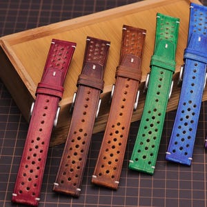 Puede incluir: Una colecci&oacute;n de correas de reloj de cuero de colores rojo, marr&oacute;n, verde, azul y morado. Cada correa presenta un dise&ntilde;o perforado y una hebilla plateada. Las correas est&aacute;n dispuestas sobre una bandeja de madera, mostrando una variedad de tonos y estilos.