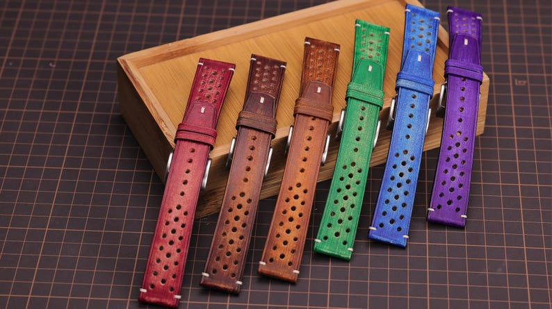 Puede incluir: Una colecci&oacute;n de correas de reloj de cuero de colores rojo, marr&oacute;n, verde, azul y morado. Cada correa presenta un dise&ntilde;o perforado y una hebilla plateada. Las correas est&aacute;n dispuestas sobre una bandeja de madera, mostrando una variedad de tonos y estilos.
