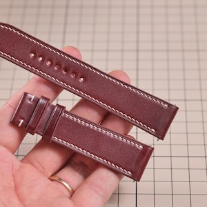 May include: Burgundy leather watch strap with white stitching. The strap has a classic design with a buckle and several holes for adjustment. The leather appears to be high-quality and durable, suitable for various watch styles.