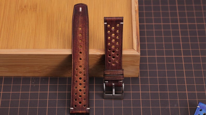 Puede incluir: Dos correas de reloj de cuero marr&oacute;n. Una es larga con peque&ntilde;os agujeros, la otra es m&aacute;s corta con agujeros m&aacute;s grandes. Ambas son de color marr&oacute;n oscuro y est&aacute;n hechas de cuero. Se muestran sobre una superficie de madera, con un patr&oacute;n de cuadr&iacute;cula en el fondo.