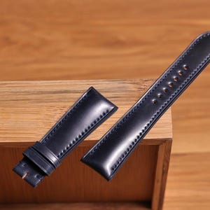 Puede incluir: Una correa de reloj de cuero azul oscuro con detalles de costura. La correa se muestra sobre una superficie de madera.