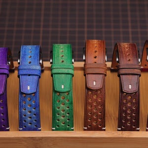 Puede incluir: Exhibici&oacute;n de seis correas de reloj de cuero de colores. Las correas son de tonos morado, azul, verde, marr&oacute;n y rojo. Cada correa tiene una serie de perforaciones circulares y est&aacute; montada en una base de madera.