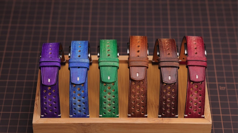 Puede incluir: Exhibici&oacute;n de seis correas de reloj de cuero de colores. Las correas son de tonos morado, azul, verde, marr&oacute;n y rojo. Cada correa tiene una serie de perforaciones circulares y est&aacute; montada en una base de madera.
