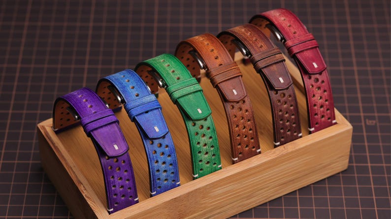 Puede incluir: Una colecci&oacute;n de correas de reloj de cuero de colores en una exhibici&oacute;n de madera. Las correas son de tonos p&uacute;rpura, azul, verde, naranja, marr&oacute;n y rojo. Cada correa tiene un dise&ntilde;o perforado y una hebilla. Las correas est&aacute;n dispuestas en fila.